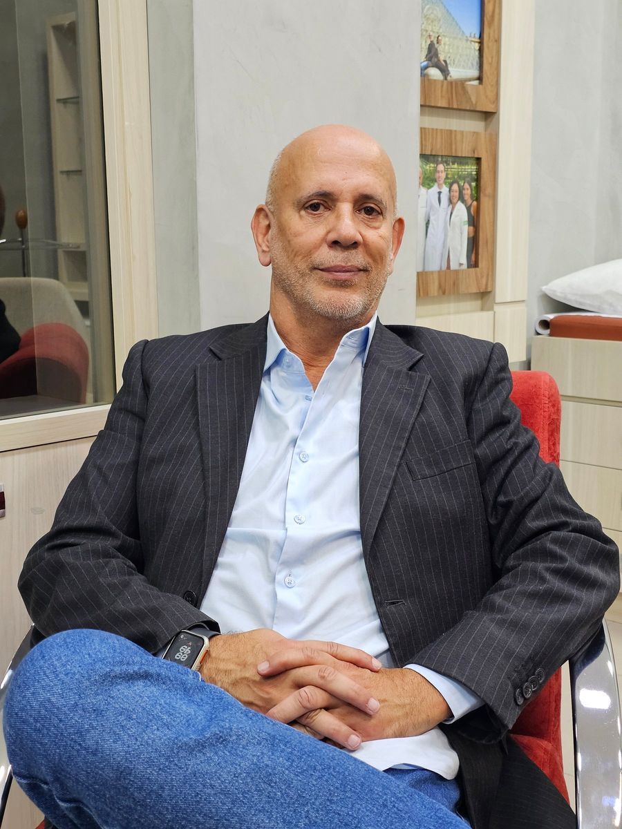 Fernando Sampaio sentado em uma poltrona com estofado terracota, dentro de um consultório com estante de madeira clara ao fundo, onde há porta retratos e uma prateleira de vidro.